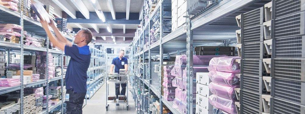 Two warehouse employees handling and transporting industrial electronic equipment stored on shelving.